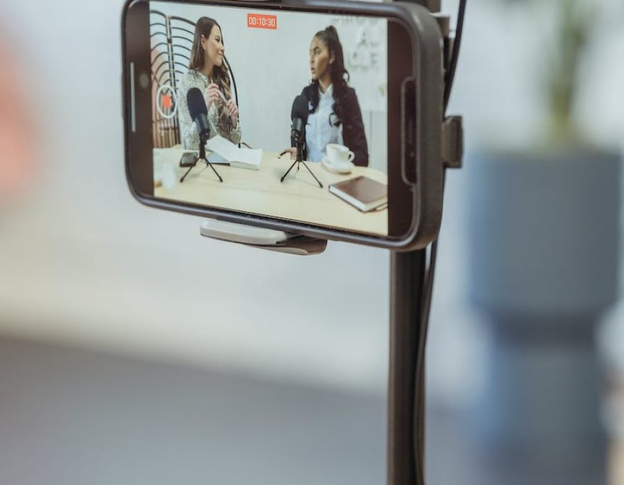 Young diverse women sitting at table with microphones and gadgets and talking while recording podcast on smartphone placed on tripod in studio